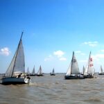 Mehrere Segelboote mit weißen Segeln liegen auf einer großen Wasserfläche unter einem klaren blauen Himmel mit vereinzelten Wolken.