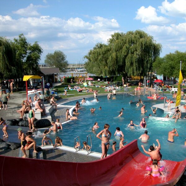 Öffentliches Freibad mit vielen Menschen, die an einem sonnigen Tag schwimmen, spielen und sich entspannen; Kinder benutzen eine rote Wasserrutsche im Vordergrund und eine Grasfläche im Hintergrund.