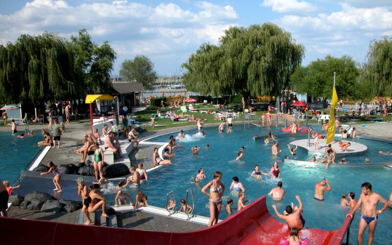 Öffentliches Freibad mit vielen Menschen, die an einem sonnigen Tag schwimmen, spielen und sich entspannen; Kinder benutzen eine rote Wasserrutsche im Vordergrund und eine Grasfläche im Hintergrund.