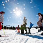 Eine Gruppe von Menschen steht in Winterkleidung und mit Skistöcken auf einer verschneiten Skipiste im hellen Sonnenlicht, über der ein strahlend blauer Himmel liegt.