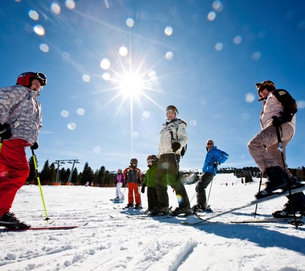 Eine Gruppe von Menschen steht in Winterkleidung und mit Skistöcken auf einer verschneiten Skipiste im hellen Sonnenlicht, über der ein strahlend blauer Himmel liegt.