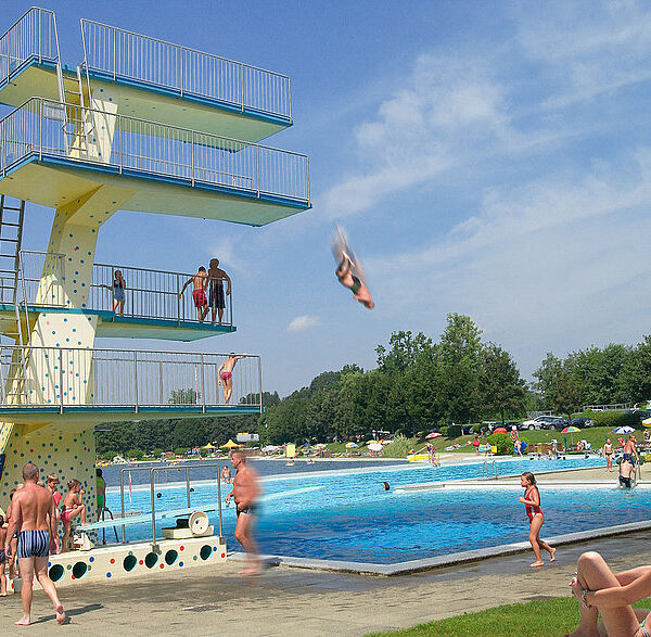 Öffentliches Freibad mit einem hohen Sprungturm; mehrere Personen schwimmen, sonnen sich, und eine Person springt in der Luft ins Wasser. Bäume und Sonnenschirme sind im Hintergrund zu sehen.