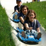 Drei Kinder fahren an einem sonnigen Tag mit einzelnen blauen Wagen eine Rodelbahn im Freien hinunter, umgeben von grünem Gras und gelben Blumen.