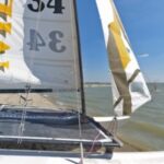 Nahaufnahme eines kleinen Segelboots mit der Nummer 34 auf dem Segel, das an einem Kiesstrand neben ruhigem Wasser unter einem klaren blauen Himmel liegt.