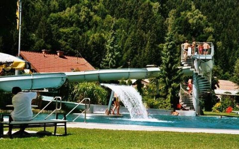 Menschen stehen auf einer Wendeltreppe und warten auf eine Wasserrutsche in einem von Bäumen und Häusern umgebenen Freibad, wobei eine Person auf einer Bank im Vordergrund sitzt.