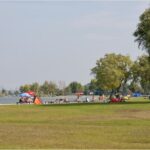 Menschen entspannen sich auf Bänken und unter Sonnenschirmen in einem grasbewachsenen Park an einem See, mit Bäumen und Segelbooten im Hintergrund.