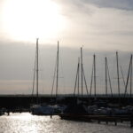 In einem Jachthafen liegen Segelboote vor Anker, deren hohe Masten sich gegen den wolkenverhangenen Himmel abzeichnen, während die Sonne tief am Horizont steht.