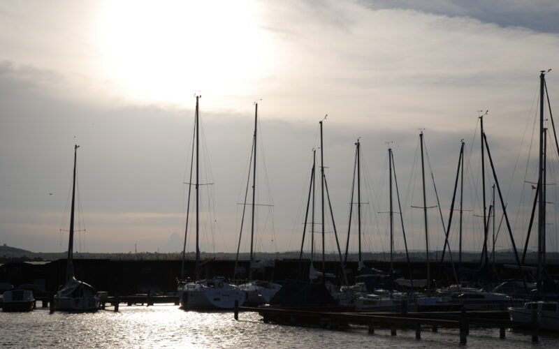 In einem Jachthafen liegen Segelboote vor Anker, deren hohe Masten sich gegen den wolkenverhangenen Himmel abzeichnen, während die Sonne tief am Horizont steht.