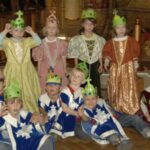 Eine Gruppe kleiner Kinder in mittelalterlichen Kostümen und mit Papierkronen posiert in einem Raum, wobei einige stehen und andere auf dem Boden sitzen.