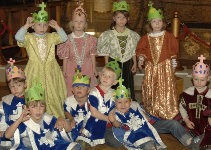 Eine Gruppe kleiner Kinder in mittelalterlichen Kostümen und mit Papierkronen posiert in einem Raum, wobei einige stehen und andere auf dem Boden sitzen.