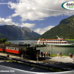 Ein historischer Zug parkt an einem Bahnhof am Seeufer, in dessen Nähe sich Menschen aufhalten; ein großes weiß-rotes Boot liegt auf dem grünen See vor Anker, umgeben von Bergen unter einem teilweise bewölkten Himmel.