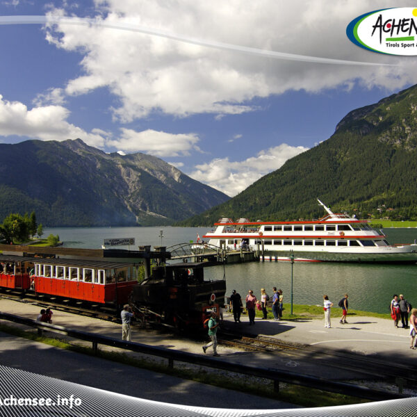 Ein historischer Zug parkt an einem Bahnhof am Seeufer, in dessen Nähe sich Menschen aufhalten; ein großes weiß-rotes Boot liegt auf dem grünen See vor Anker, umgeben von Bergen unter einem teilweise bewölkten Himmel.