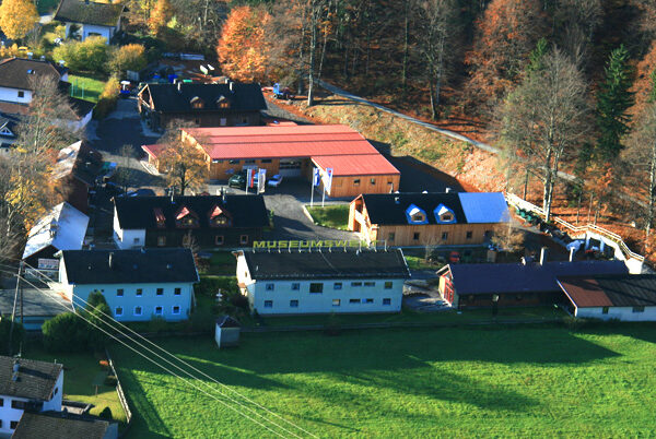 Luftaufnahme eines kleinen Museumskomplexes mit mehreren Gebäuden, umgeben von Bäumen und Grasflächen, am Rande eines Waldes gelegen.