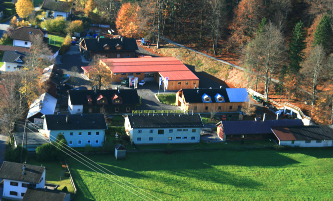 Luftaufnahme eines kleinen Museumskomplexes mit mehreren Gebäuden, umgeben von Bäumen und Grasflächen, am Rande eines Waldes gelegen.