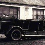 Schwarz-Weiß-Foto eines Autos aus dem frühen 20. Jahrhundert, das vor einem Gebäude mit verschlossenen Fenstern geparkt ist. Auf der Rückseite ist ein Ersatzreifen montiert.