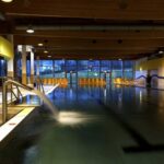 Hallenbad mit Wasserspeier, Metallgeländer und Reihen von gelben Liegestühlen entlang großer Fenster, die Tageslicht hereinlassen.