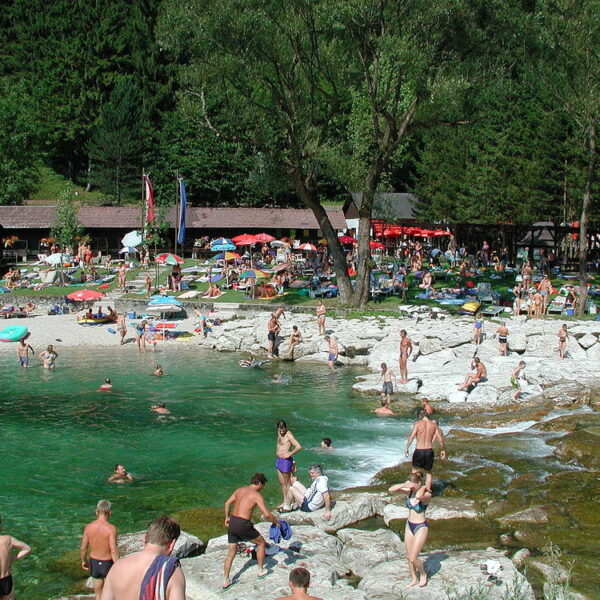Die Menschen schwimmen, sonnen sich und entspannen sich in der Nähe eines felsigen Ufers und klaren grünen Wassers, mit Bäumen und einer Grasfläche mit Sonnenschirmen und Gebäuden im Hintergrund.
