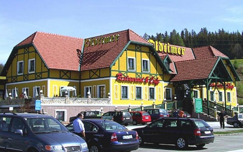 Ein gelb-grünes Restaurant mit roten Dachziegeln, Außentreppen und parkenden Autos auf dem Parkplatz an einem klaren Tag, umgeben von Grün und Bäumen.