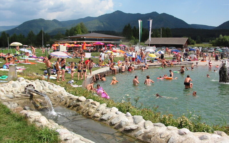 Ein überfüllter Freibadbereich mit Menschen, die sich sonnen, sitzen und schwimmen, umgeben von Gras, Sonnenschirmen und Bergen im Hintergrund.