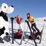 Vier Personen posieren auf einer verschneiten Piste mit einer Person in einem Hundemaskottchen-Kostüm; zwei stehen, während zwei mit V-förmig angeordneten Skiern auf dem Schnee sitzen.