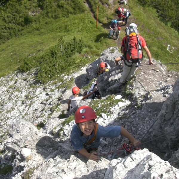 Eine Gruppe von Kletterern mit Helmen und Klettergurten steigt an einem sonnigen Tag mit Hilfe von Sicherungsseilen einen felsigen, steilen Hang hinauf.