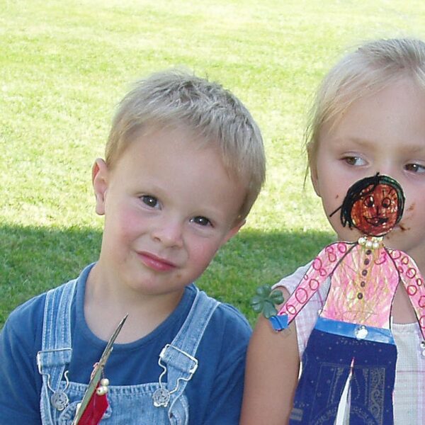 Ein kleiner Junge in einem Overall und ein kleines Mädchen mit einer handgefertigten Papierpuppe stehen zusammen auf einer Wiese.