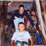 Fünf Kinder posieren gemeinsam auf einer Holztreppe in einem Tunnel oder einer Höhle, die von Holzbalken umgeben zu sein scheint.