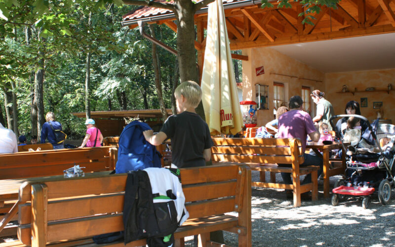 Menschen, darunter auch Kinder, sitzen und stehen um hölzerne Bänke und Tische im Freien unter einem Baum in der Nähe eines Gebäudes mit einem roten Ziegeldach. Das Sonnenlicht fällt durch die Blätter.
