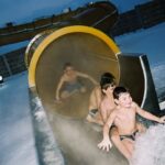 Drei Kinder in Badeanzügen verlassen eine Wasserrutsche in einem schneebedeckten Außenbereich, während ein anderes Kind noch in der Rutsche steht.