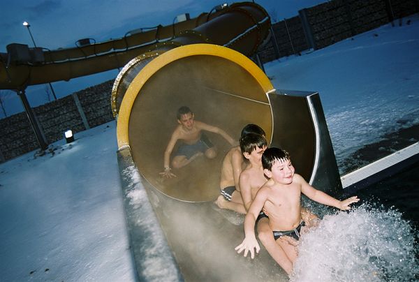Drei Kinder in Badeanzügen verlassen eine Wasserrutsche in einem schneebedeckten Außenbereich, während ein anderes Kind noch in der Rutsche steht.