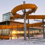 Eine große gelbe Wasserrutsche im Freien windet sich um Metallstützen neben einem modernen Glasgebäude, während der Boden mit Schnee bedeckt ist und die Sonne sich in den Fenstern spiegelt.