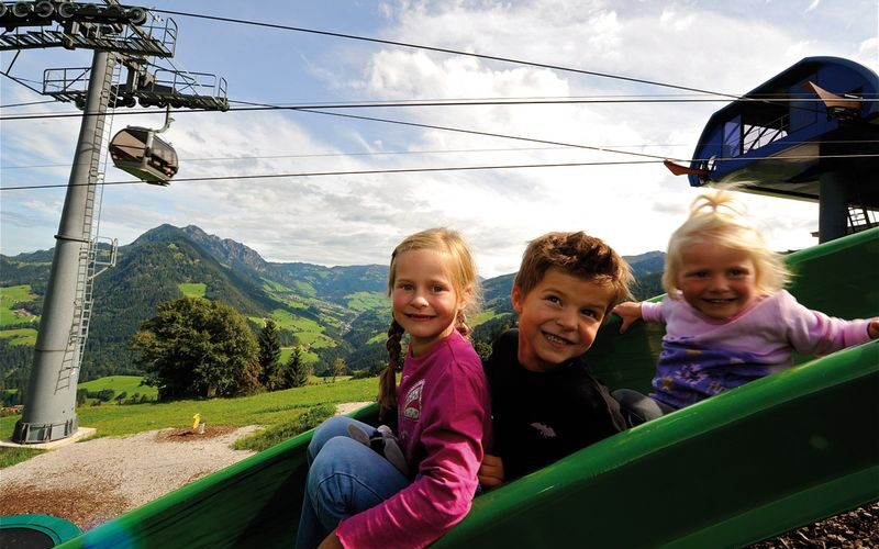 Drei kleine Kinder rutschen auf einer grünen Rutsche im Freien mit Bergen, Bäumen und einer Seilbahn im Hintergrund unter einem teilweise bewölkten Himmel hinunter.