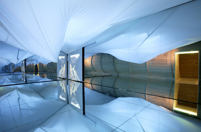 Raum mit verspiegeltem Boden, weißer Stoffdecke, Holzwand mit Tür und einem vertikalen Landschaftsfoto, das sich im Spiegel spiegelt.