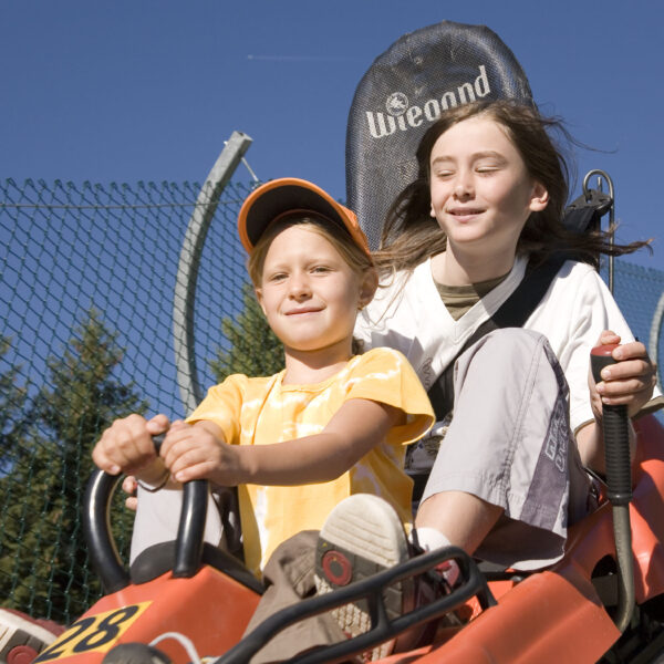 Zwei Kinder fahren an einem sonnigen Tag im Freien in einem roten Achterbahnwagen, im Hintergrund sind Bäume und ein blauer Himmel zu sehen.