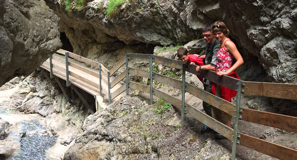 Eine Familie steht auf einem Holzsteg mit Metallgeländer, der entlang einer felsigen Schlucht oberhalb eines Baches gebaut und von steilen Felswänden umgeben ist.