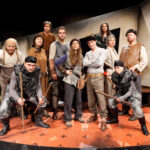 Eine Gruppe von Menschen in mittelalterlichen Kostümen posiert auf einer Theaterbühne mit Requisiten und bereitet sich wahrscheinlich auf oder nach einer Theateraufführung vor.