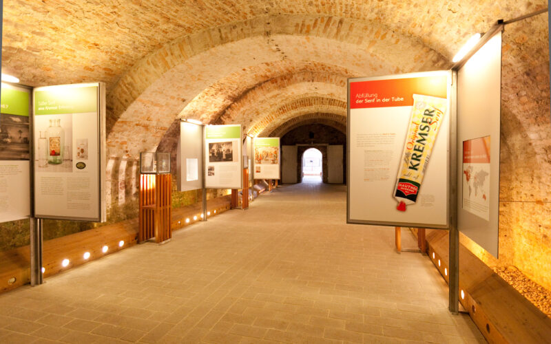 In einem Flur mit Ziegelgewölbe sind Informationstafeln und Bilder zu sehen, darunter das große Bild einer Senftube, in einem Museum oder einer Ausstellung.