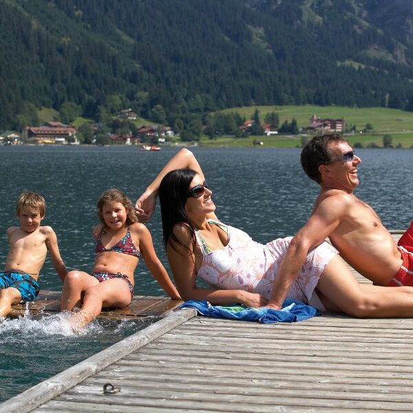 Eine vierköpfige Familie entspannt sich auf einem hölzernen Steg an einem See; zwei Kinder sitzen mit den Füßen im Wasser, während zwei Erwachsene in Badeanzügen vor dem Hintergrund der Berge und Bäume faulenzen.