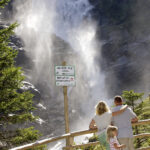 Eine dreiköpfige Familie steht an einem Holzzaun und blickt auf einen großen Wasserfall; ein Schild warnt die Besucher, aus Sicherheitsgründen hinter der Absperrung zu bleiben.