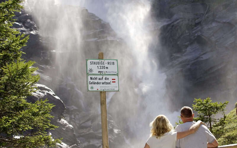 Eine dreiköpfige Familie steht an einem Holzzaun und blickt auf einen großen Wasserfall; ein Schild warnt die Besucher, aus Sicherheitsgründen hinter der Absperrung zu bleiben.