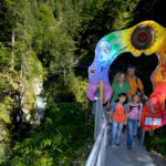 Vier Personen stehen auf einem Metallsteg in einer bewaldeten Schlucht und gehen unter einem farbenfrohen, kunstvollen Torbogen mit Text und Symbolen hindurch.