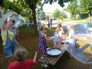 An einem sonnigen Tag spielen Kinder im Freien unter Bäumen und machen große Seifenblasen mit Stäben und Schalen mit Seifenblasenlösung.