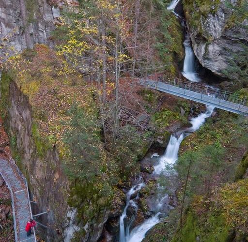 Erhöhte Metallstege überspannen eine enge Schlucht mit Wasserfällen, umgeben von Bäumen und moosbewachsenen Felsen; eine Person in einer roten Jacke geht auf dem Weg.