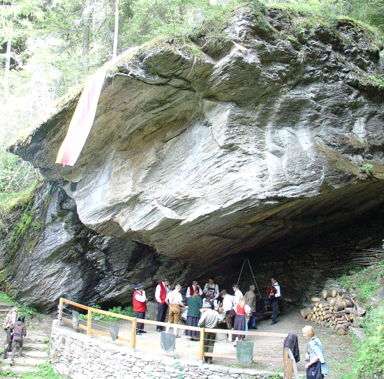 Eine Gruppe von Menschen in traditioneller Kleidung versammelt sich unter einem großen Felsüberhang in einem Waldgebiet, in dessen Nähe Baumstämme und aufgeschichtete Steine liegen.
