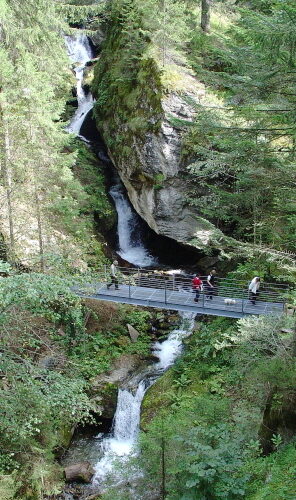 Eine Gruppe von Menschen überquert einen Metallsteg über einen Bach mit einem Wasserfall, der von dichtem Wald umgeben ist.