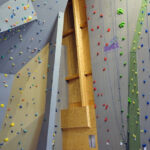 Kletterhalle mit hohen künstlichen Kletterwänden, mehrfarbigen Griffen, Seilen und einem schwarzen Metallgeländer mit einem einzelnen Stuhl im Vordergrund.