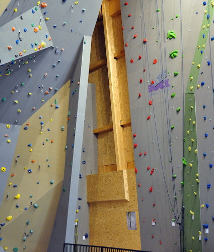 Kletterhalle mit hohen künstlichen Kletterwänden, mehrfarbigen Griffen, Seilen und einem schwarzen Metallgeländer mit einem einzelnen Stuhl im Vordergrund.