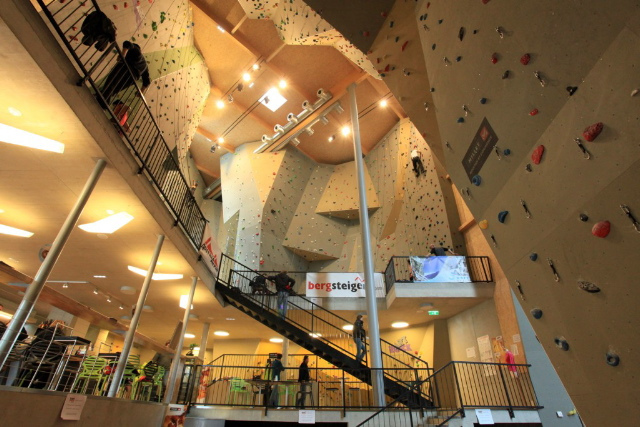 Indoor-Kletterhalle mit hohen Kletterwänden, künstlichen Griffen, Treppen, Geländern und kletternden und gehenden Menschen in einer geräumigen, gut beleuchteten Anlage.