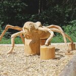 Große Holzskulptur einer Ameise mit sechs Beinen, die auf Holzspänen in einem bewaldeten Außenbereich steht.