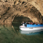Eine Gruppe von Menschen fährt in einem weißen Boot mit blauen Sitzen durch eine unterirdische Höhle, die mit klarem Wasser gefüllt ist.